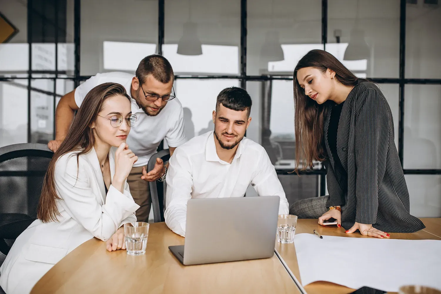 Four colleagues gathered around a laptop at a wooden table in a bright office, discussing their work
